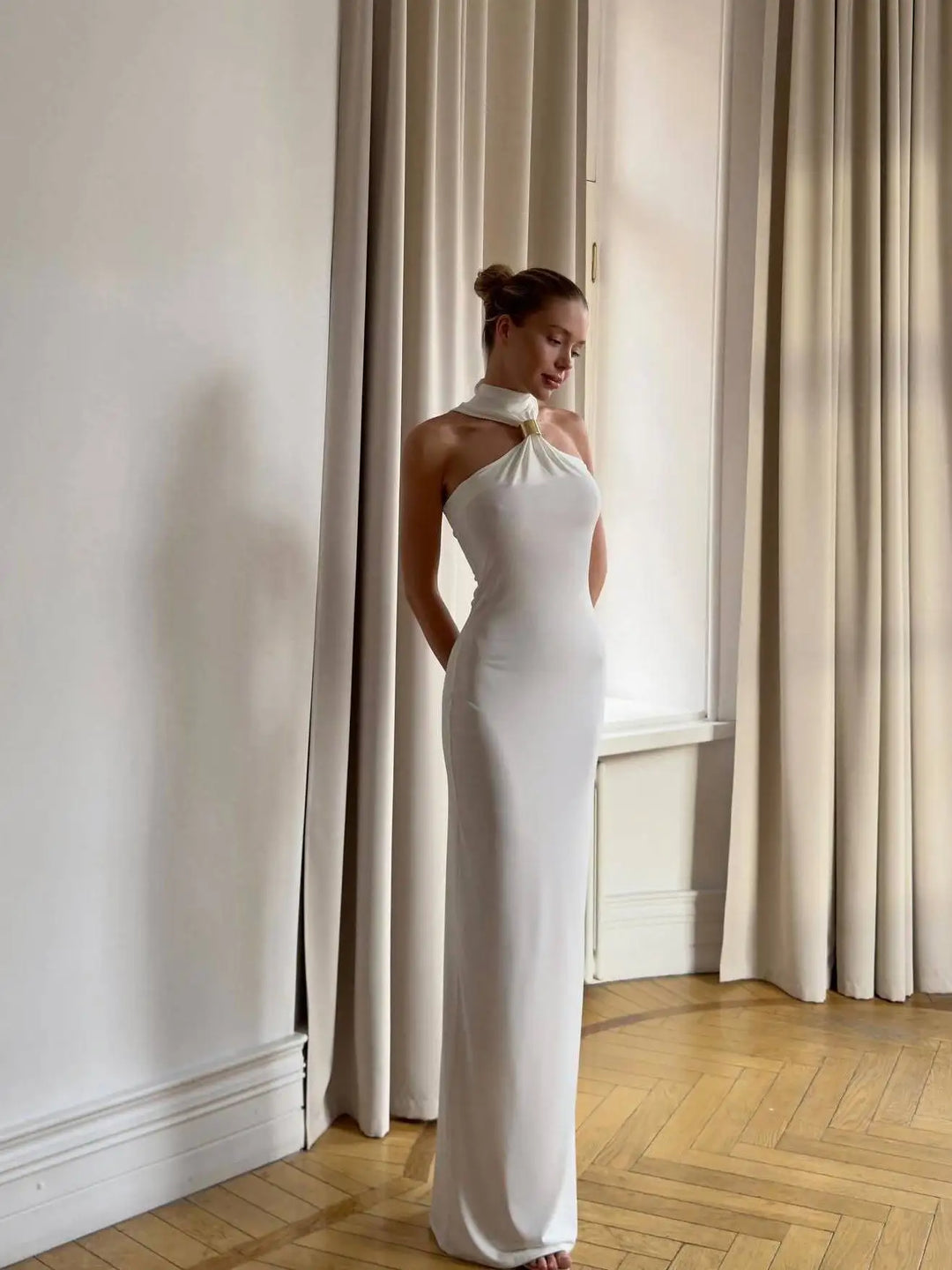 A woman in a white maxi dress with a halter neck and a backless design, standing in a room with floor-to-ceiling windows.