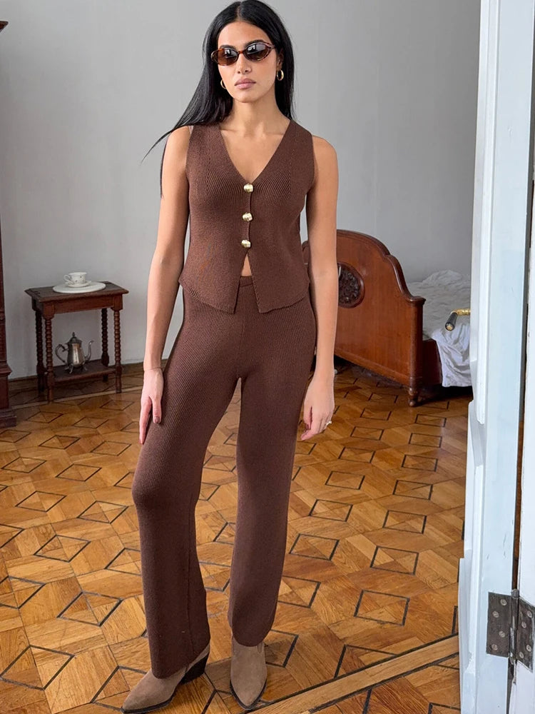 Woman wearing a brown outfit standing in a room with wooden flooring and a bed.