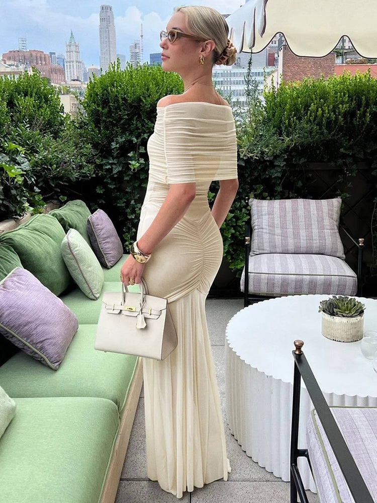 Woman in a beige off-shoulder dress standing in an outdoor setting with city skyline in the background.