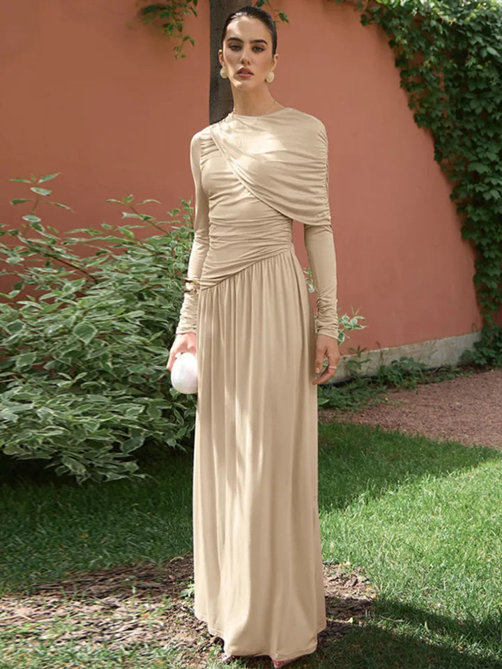 Woman in a beige long-sleeve dress standing outdoors with greenery and a pink wall in the background.