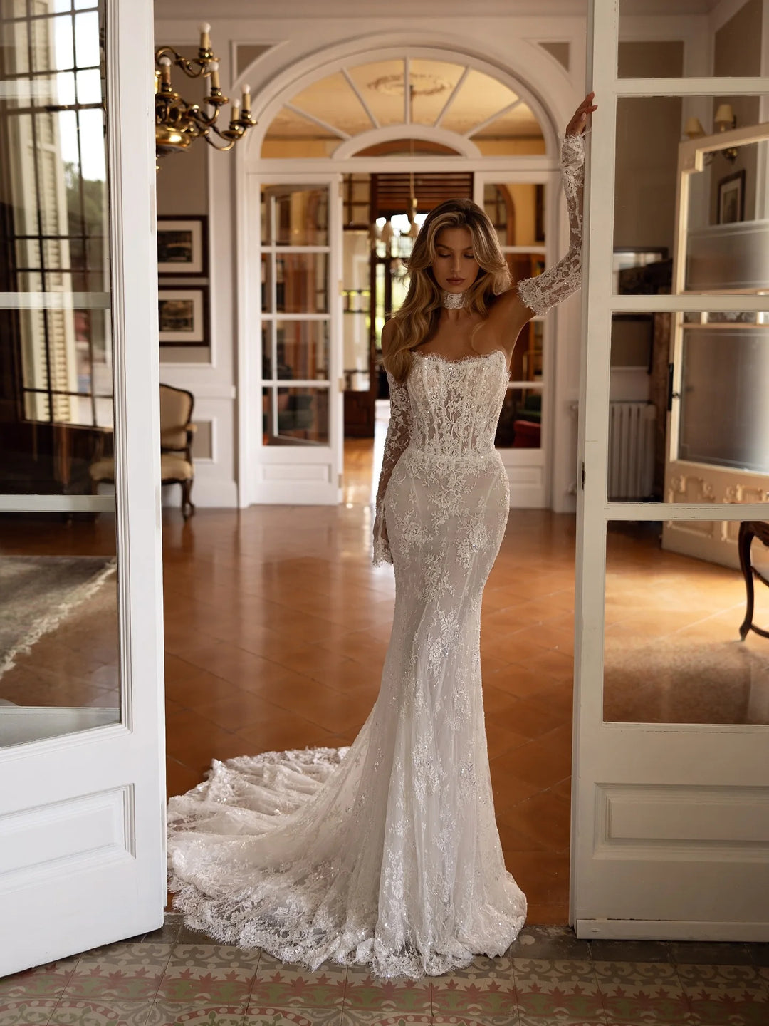 Woman in a white lace wedding dress standing in a room with large windows.