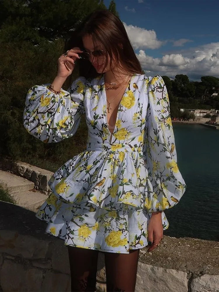 A woman standing by the water wearing a floral print mini dress with long sleeves and a deep v-neck.
