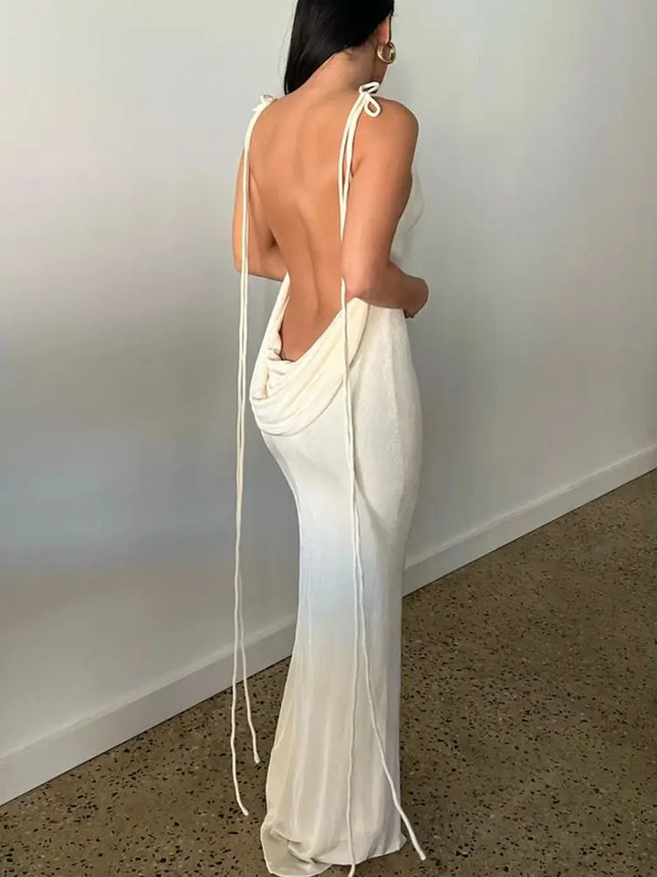 Woman wearing a white evening gown with a backless design against a plain wall.