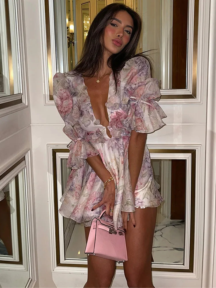 A woman standing in an elegant room wearing a floral mini dress with ruffled sleeves and a deep v-neck.