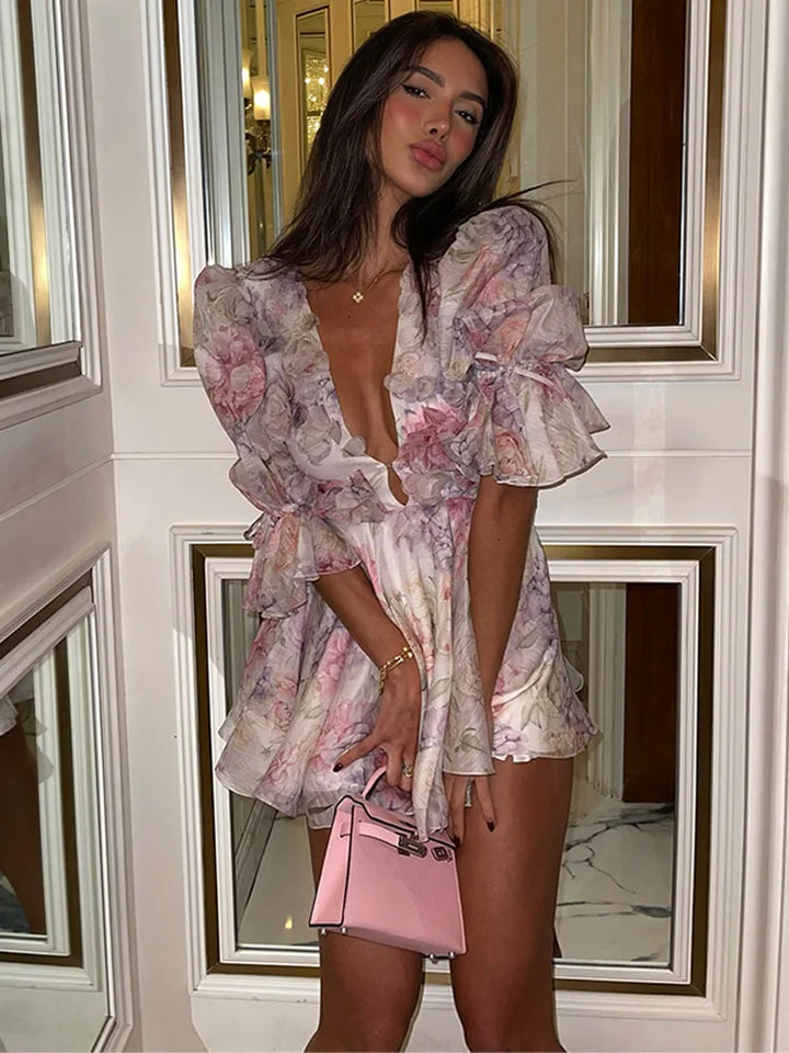 A woman standing in an elegant room wearing a floral mini dress with ruffled sleeves and a deep v-neck.
