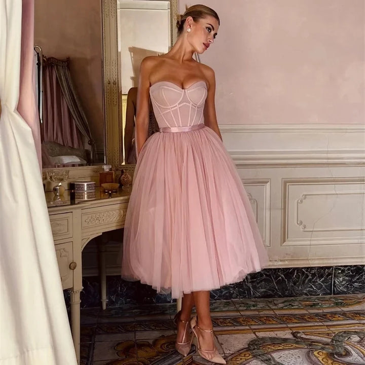 A woman standing in a room wearing a pink strapless midi dress with a sweetheart neckline and a full skirt.