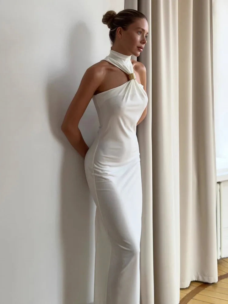 A woman in a white maxi dress with a halter neck and a backless design, standing in a room with floor-to-ceiling windows.