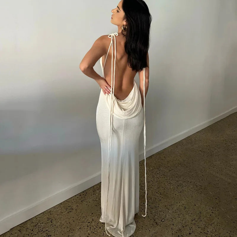 Woman wearing a white dress with a backless design, standing against a plain wall.