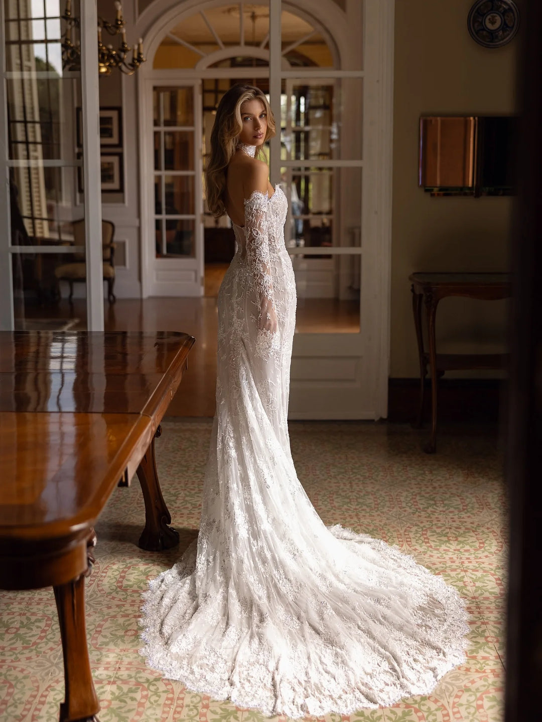 Woman in a white lace wedding dress standing in a room with wooden furniture and a large mirror.