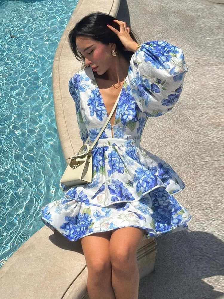A woman standing by the water wearing a floral print mini dress with long sleeves and a deep v-neck.