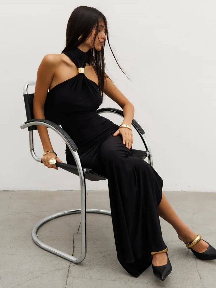 A woman in a bkack maxi dress with a halter neck and a backless design, standing in a room with floor-to-ceiling windows.