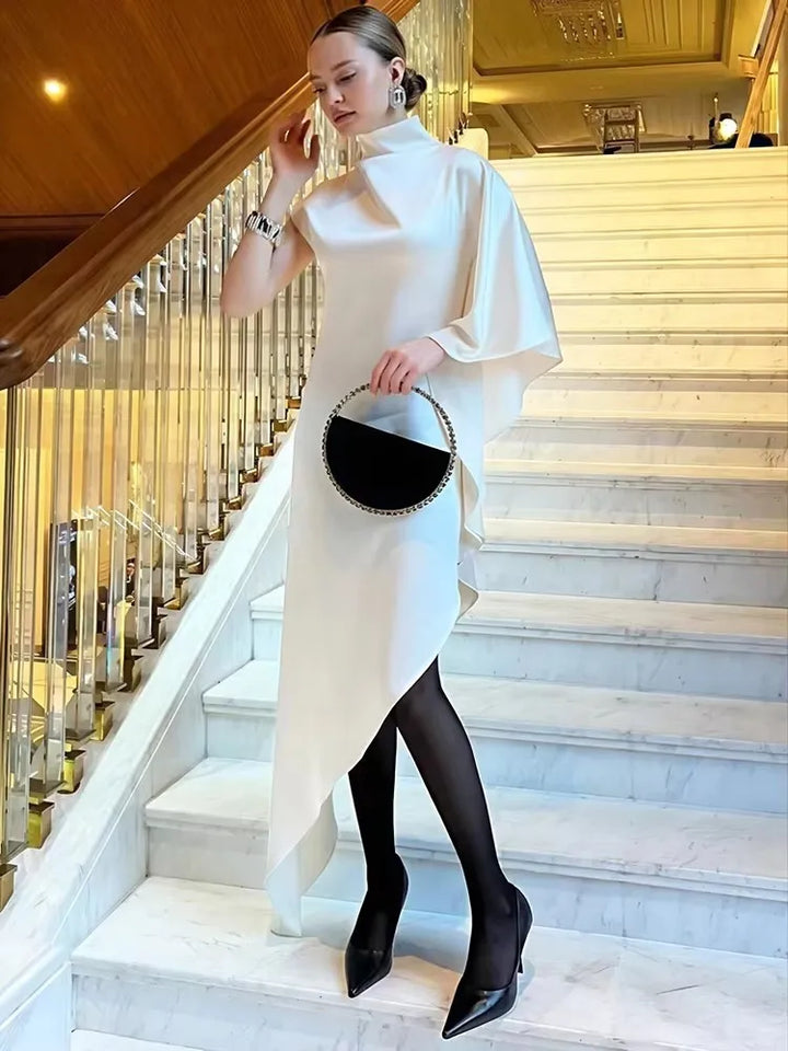 Elegant woman in a satin sleeveless Kiersten Maxi Dress with irregular hem and turtleneck, posing on a staircase.