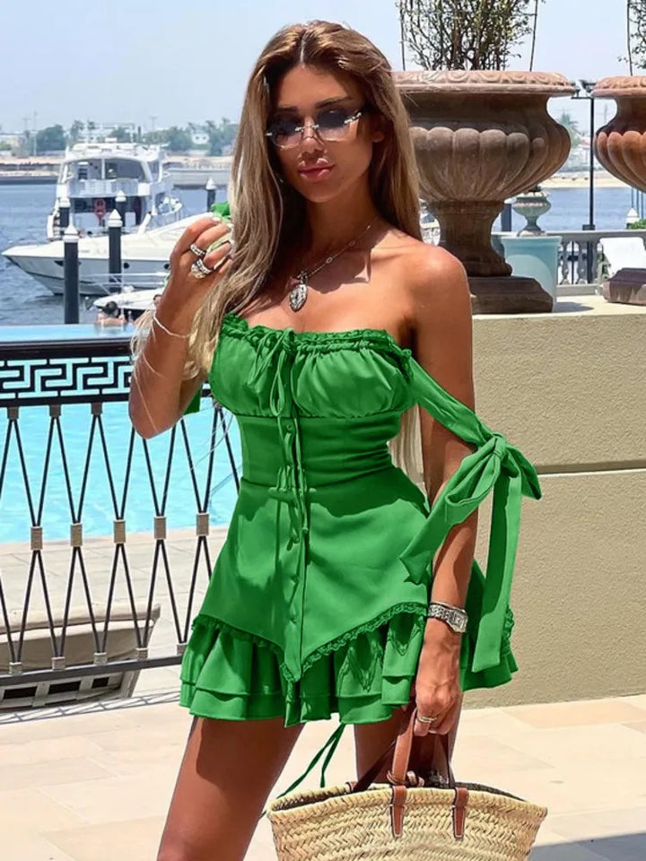 A woman standing near a body of water wearing a green sleeveless, ruffled, off-the-shoulder mini dress with lace-up back and spaghetti straps. She is also wearing sunglasses and carrying a straw bag.