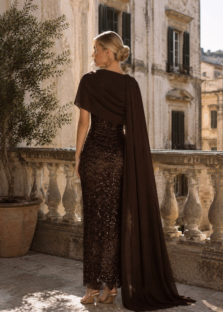 Woman in a long, dark brown dress with a cape standing in an elegant outdoor setting.