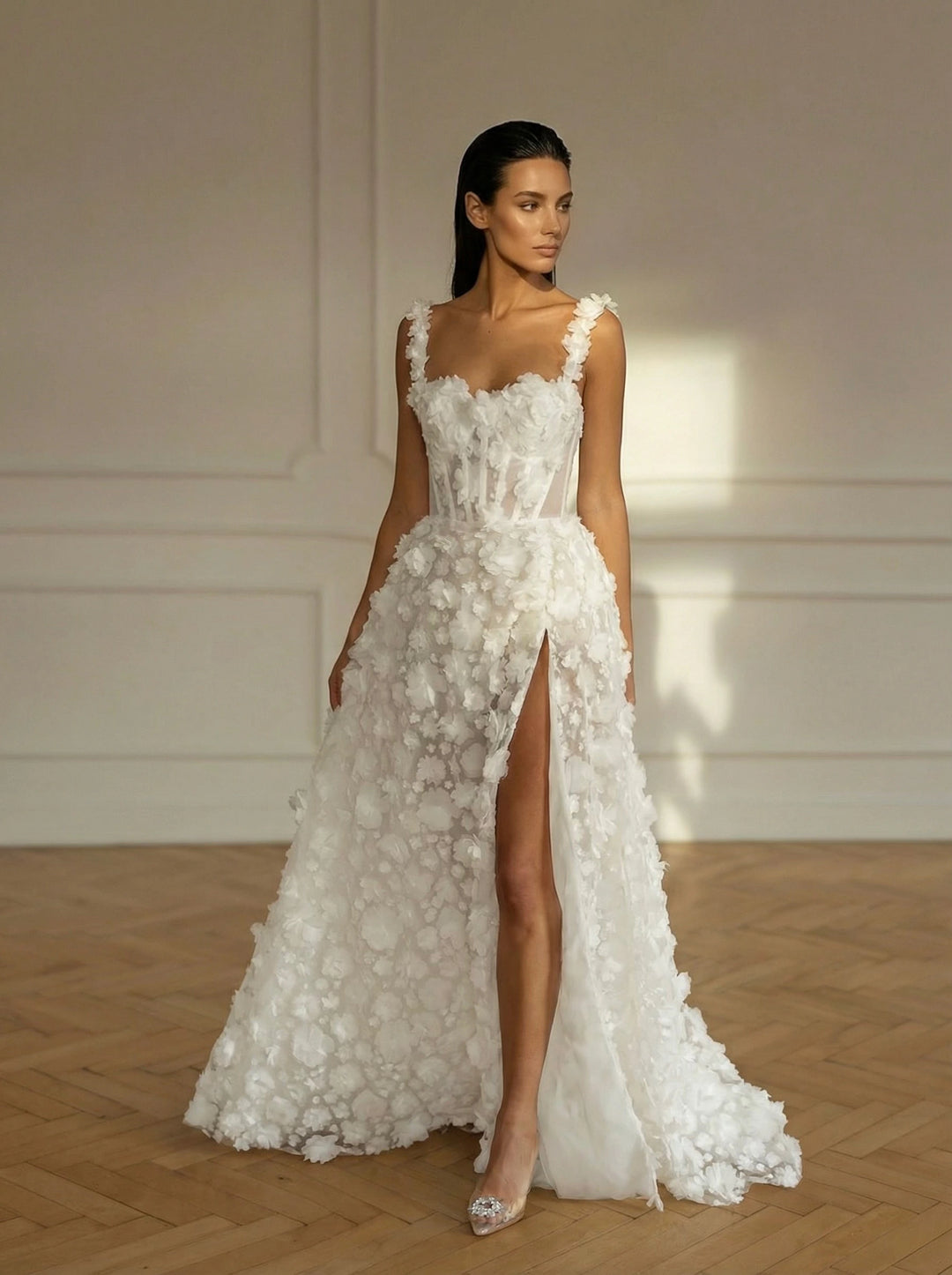 Woman wearing a white floral wedding dress with a high slit in a room with wooden flooring and white walls.