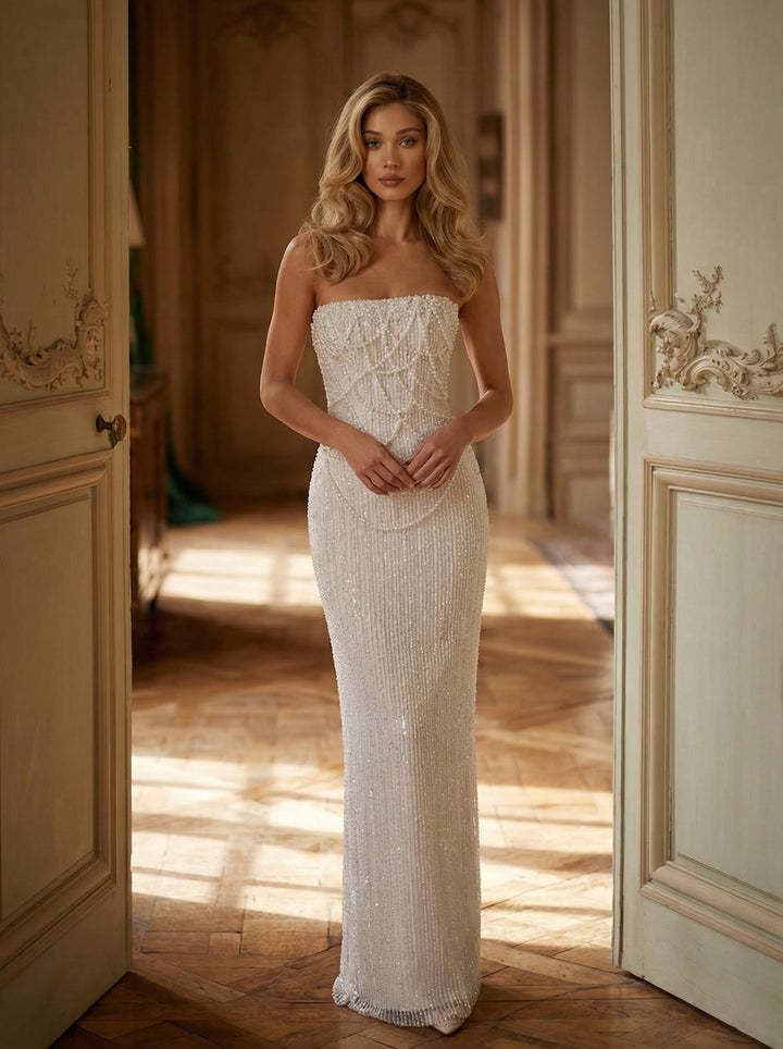 Woman in a white strapless gown standing in a sunlit room with wooden floors and decorative walls.