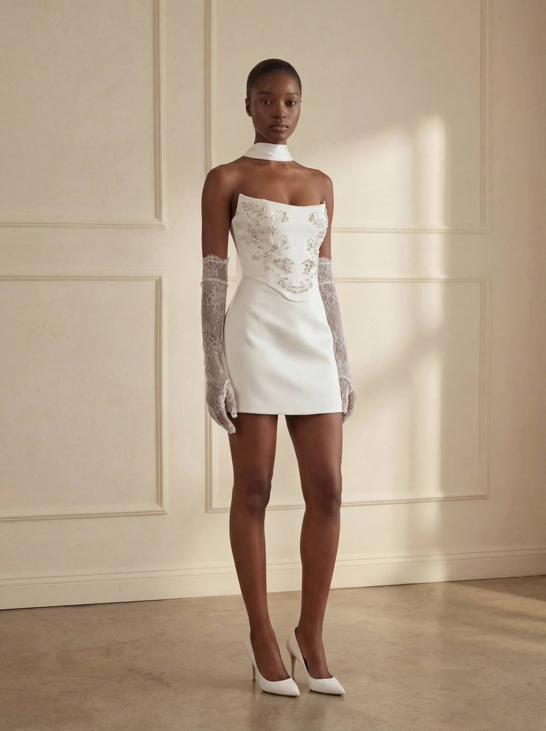 Woman wearing a white dress with lace sleeves against a beige wall.