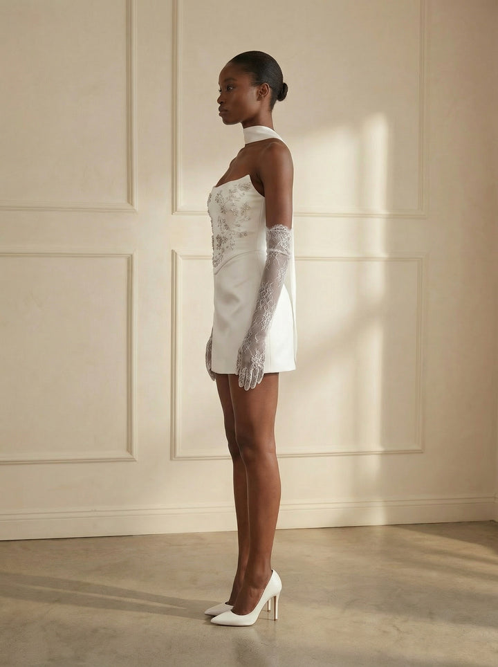 Woman wearing a white dress with lace detailing and white gloves against a beige wall.