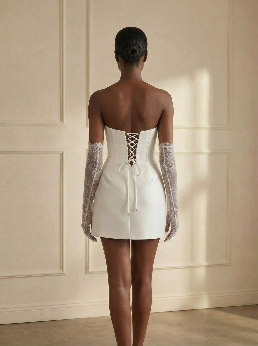 Woman wearing a white dress with lace-up back and sheer sleeves against a beige wall.