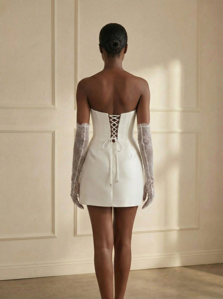 Woman wearing a white dress with lace-up back and sheer sleeves against a beige wall.