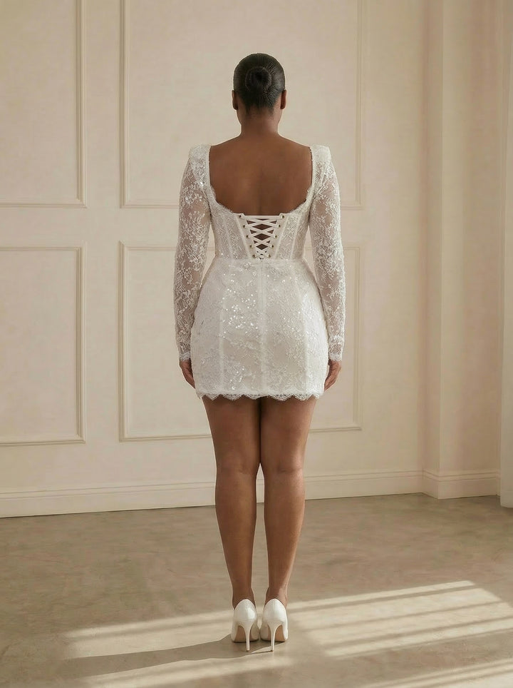 Woman wearing a white lace dress with long sleeves in a room with light-colored walls.