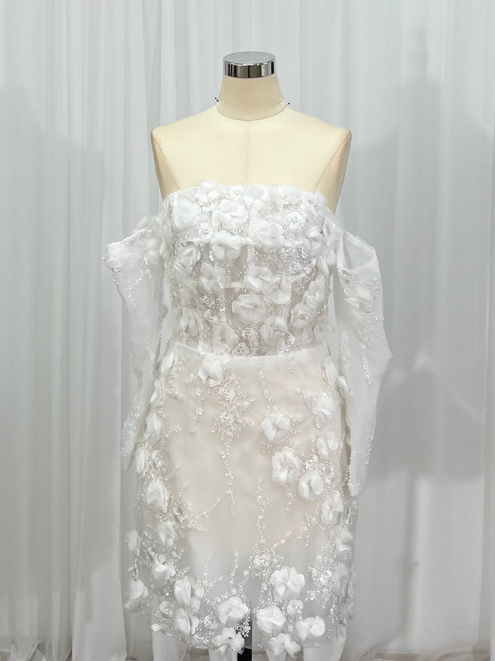 A strapless white wedding dress with 3D floral appliqués and sheer draped sleeves, displayed on a cream mannequin against a white curtain backdrop.