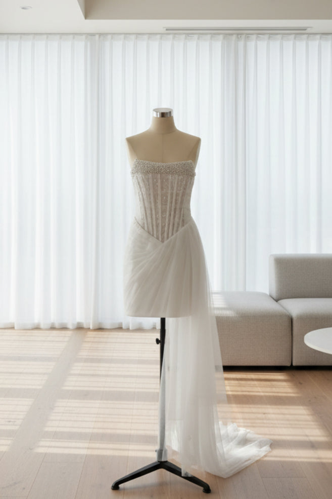 A white strapless wedding dress with a beaded corset-style bodice and a sheer tulle overskirt, displayed on a mannequin stand in front of white draped curtains.