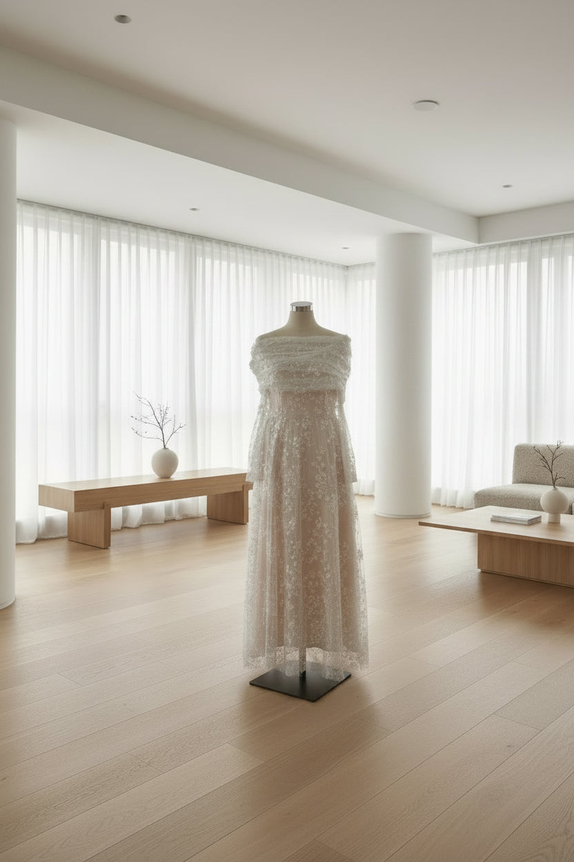 A long, heavily embellished formal gown displayed on a dress form in an indoor room with curtains, a carpeted floor, and a pinned design board on the wall.