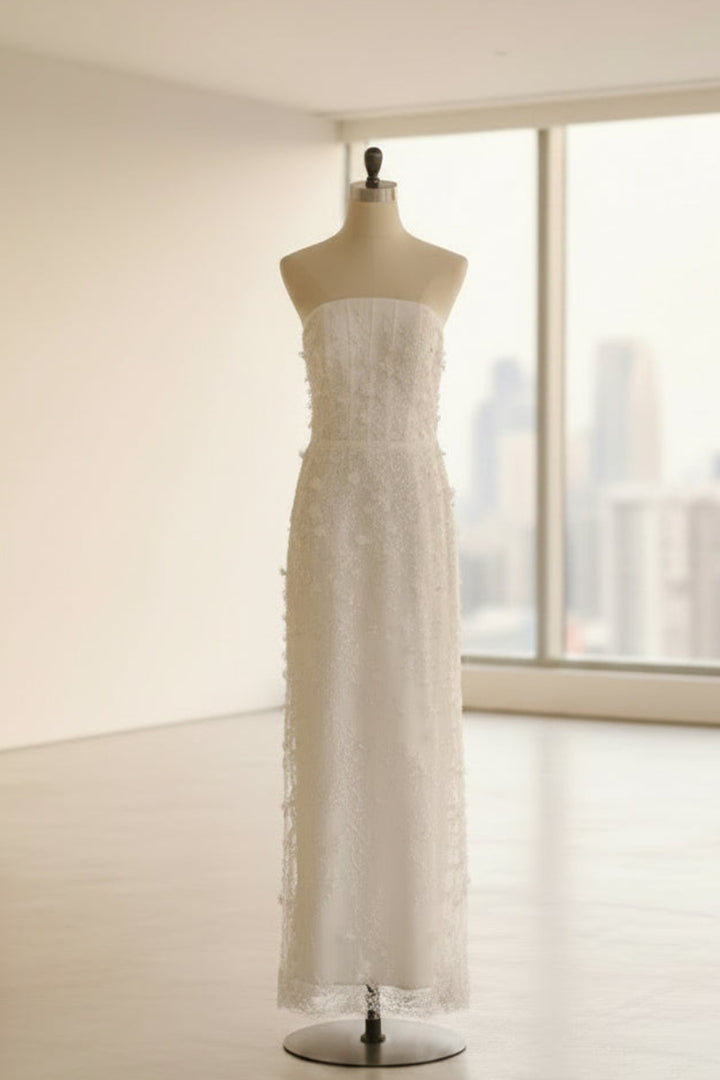 A strapless white lace gown displayed on a dress form mannequin in a bright, minimalist studio with large windows and a city skyline view.