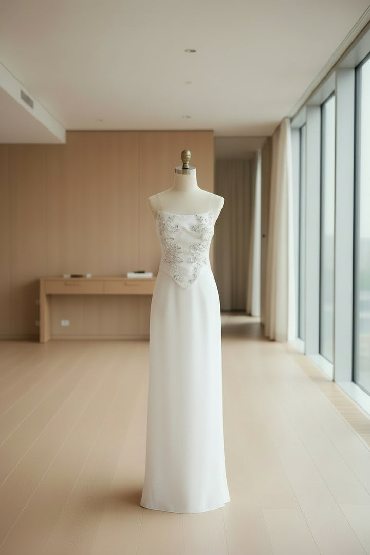 A white strapless formal gown displayed on a dress form in a sewing room. The bodice features delicate silver floral embroidery, and the skirt is a smooth, floor-length white satin-like fabric.