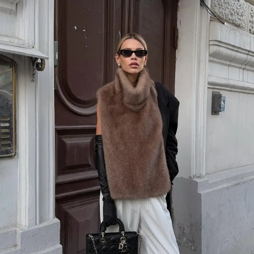 Woman wearing a brown fur vest, black sunglasses, and white pants standing in front of a building entrance.