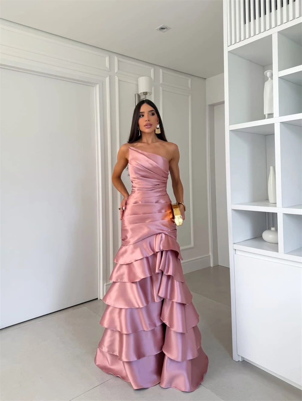 Woman in a pink one-shoulder gown standing in a room with white walls and a bookshelf.