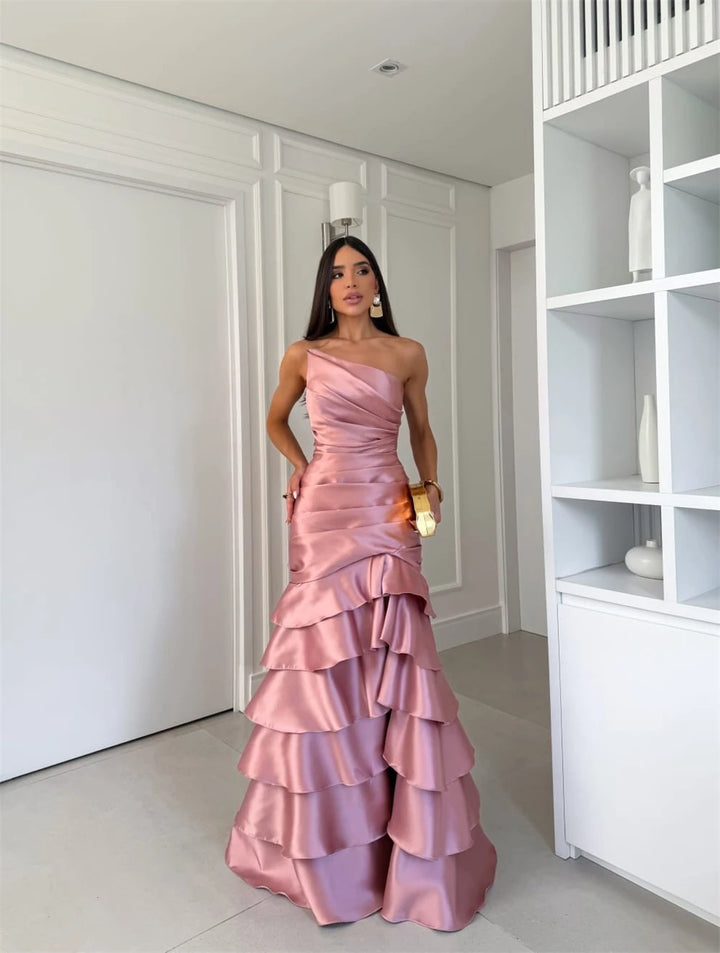 Woman in a pink one-shoulder gown standing in a room with white walls and a bookshelf.