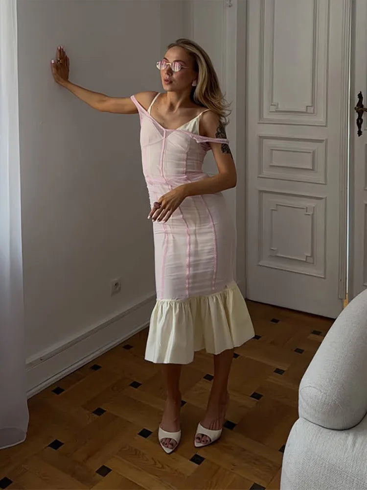 A woman wearing a light pink and white striped off-the-shoulder midi dress with a ruffled hem, standing in a room with a wooden floor and white paneled doors.