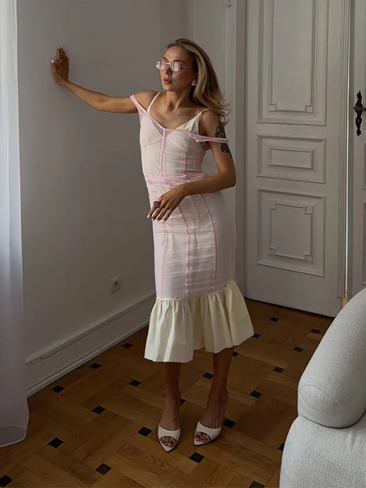A woman wearing a light pink and white striped off-the-shoulder midi dress with a ruffled hem, standing in a room with a wooden floor and white paneled doors.