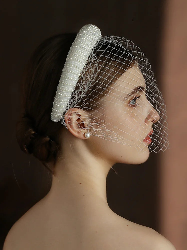 Woman wearing a white beaded headband and birdcage veil against a dark background