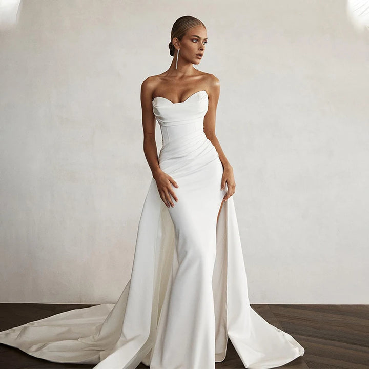 Woman wearing a strapless white wedding dress with a slit on a plain background