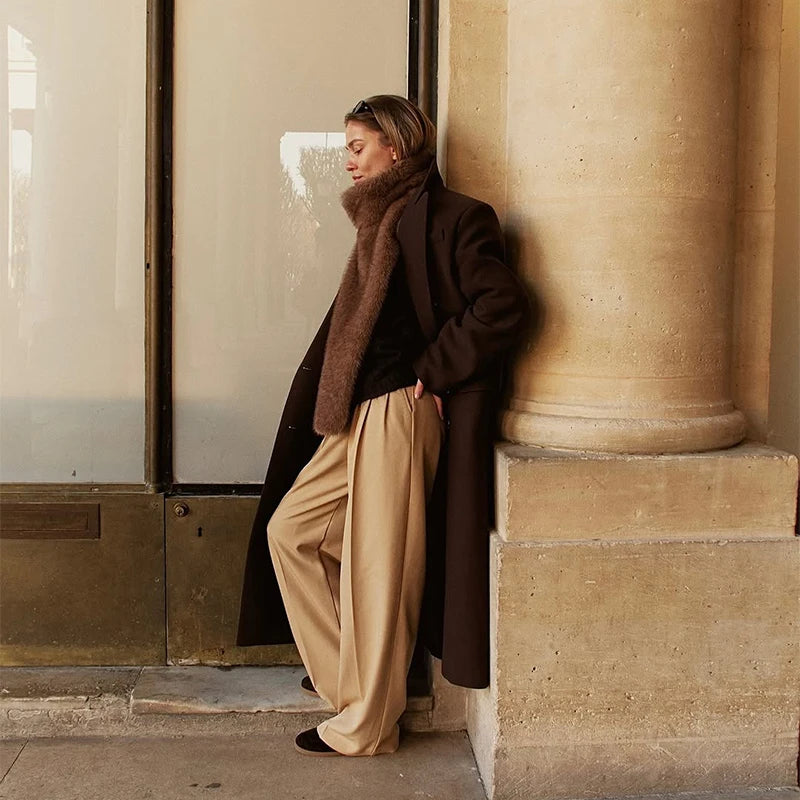 Woman leaning against a wall in a stylish outfit with a coat and scarf.