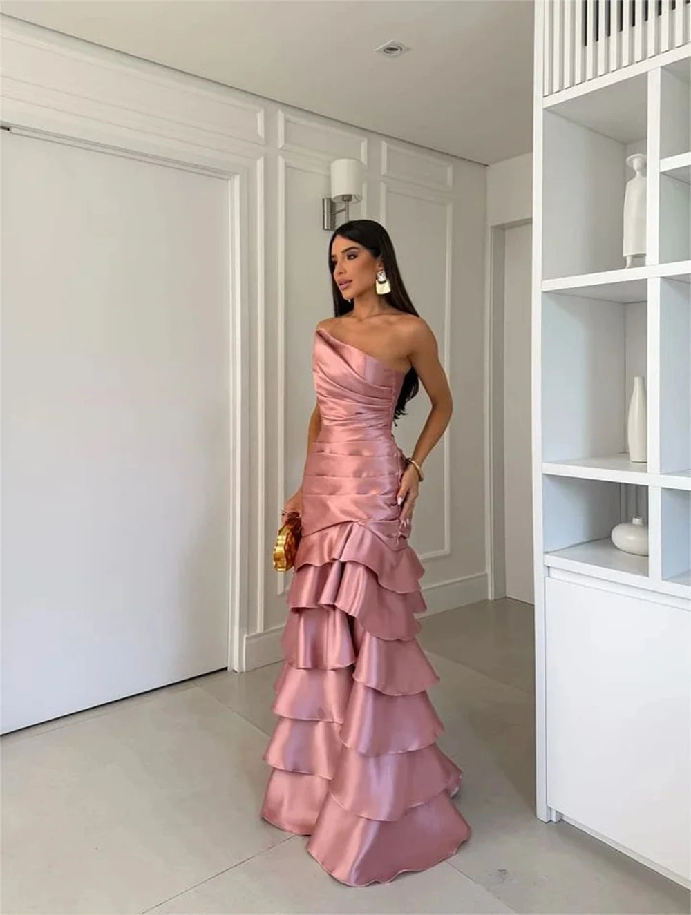 Woman in a pink one-shoulder gown standing in a room with white walls and a bookshelf.