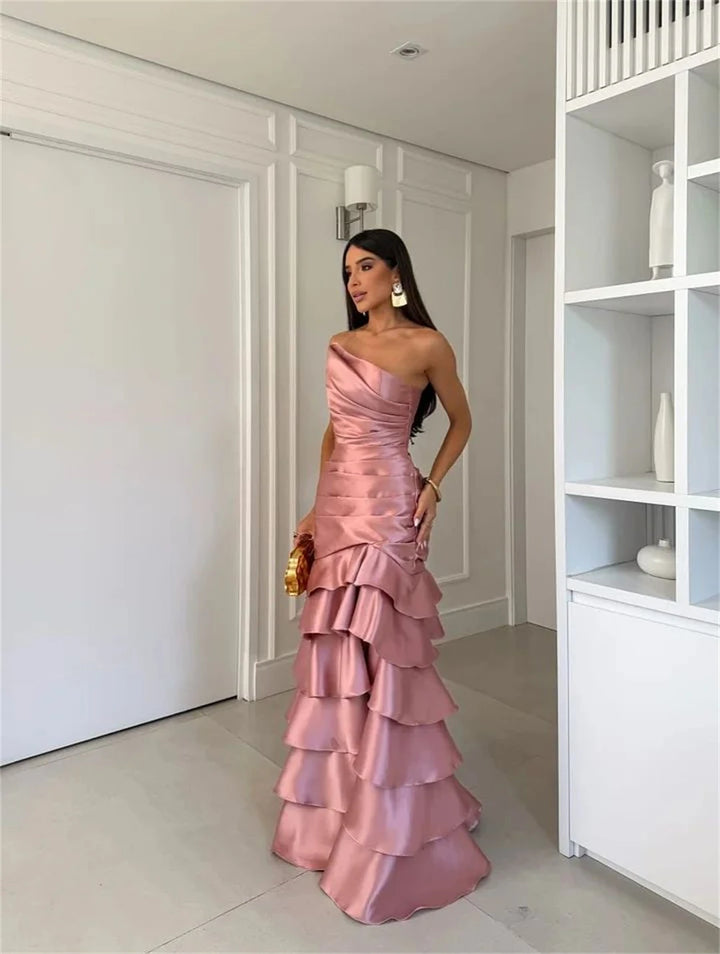 Woman in a pink one-shoulder gown standing in a room with white walls and a bookshelf.