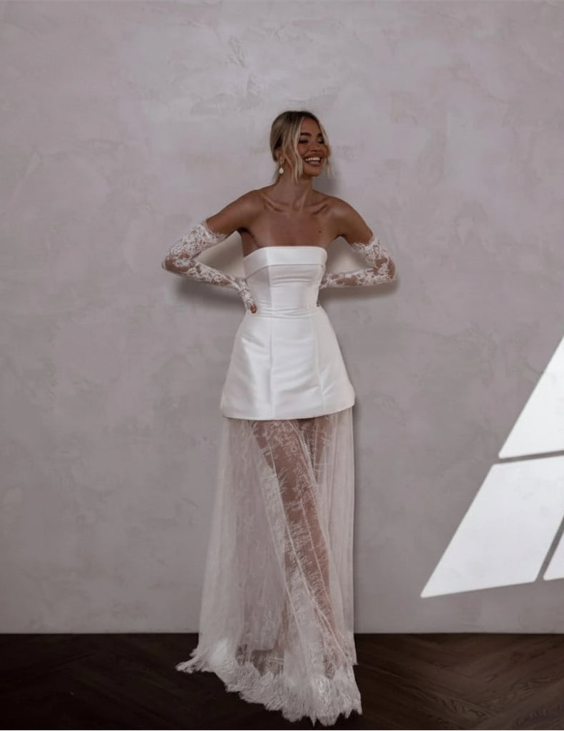 A woman models a white strapless dress with long sheer lace sleeves, a structured corset-style bodice, and a flowing lace overlay skirt, standing against a textured gray wall with a geometric window light on the right. The brand name &