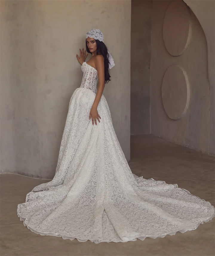 A woman modeling a strapless white lace wedding gown with a fitted bodice, full skirt, and long train in a minimalist concrete-walled interior.