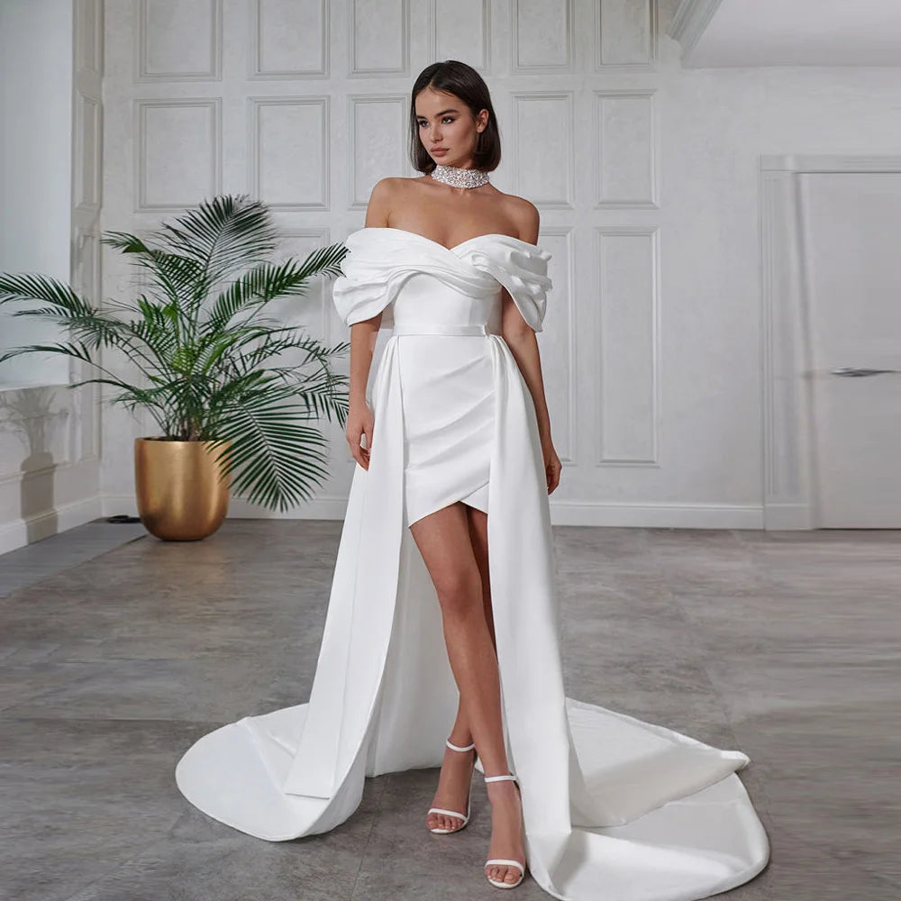 Woman in a white off-shoulder gown standing in a room with a plant and white walls.