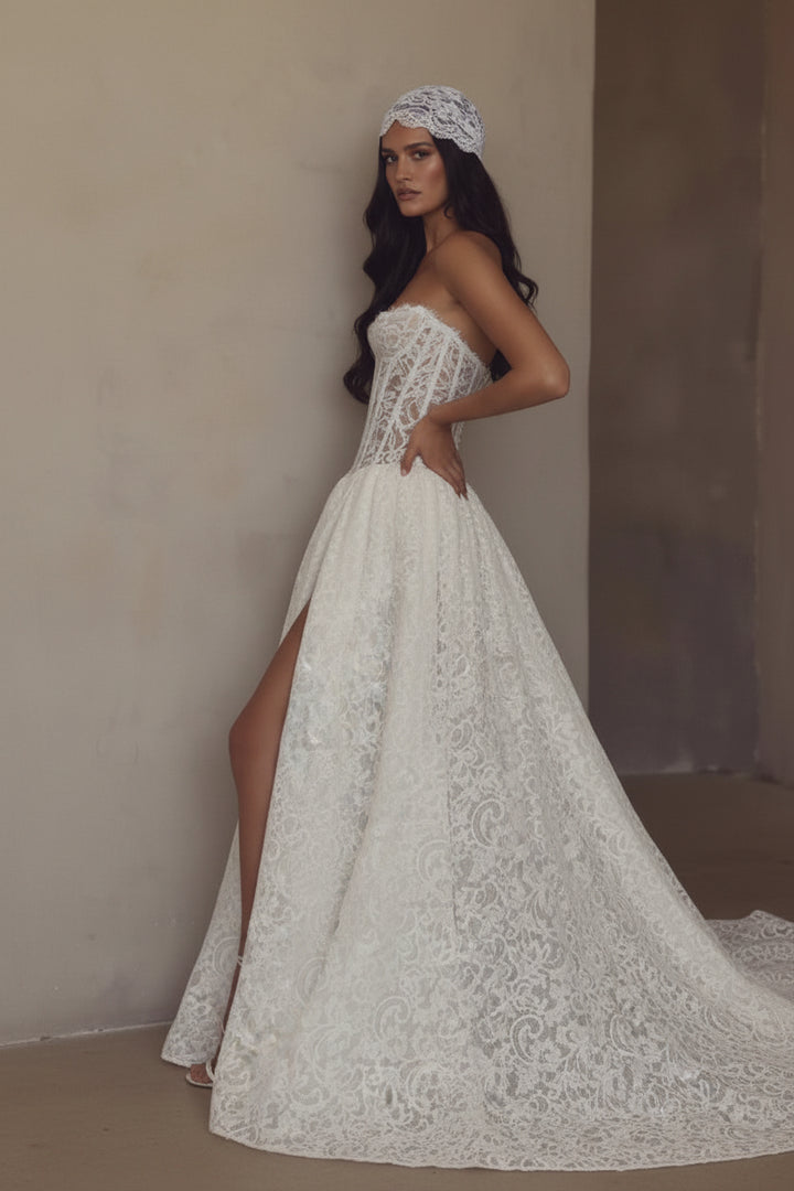 A woman models a strapless white lace wedding gown with a fitted corset-style bodice, high front slit, and long flowing train against a warm beige textured wall.