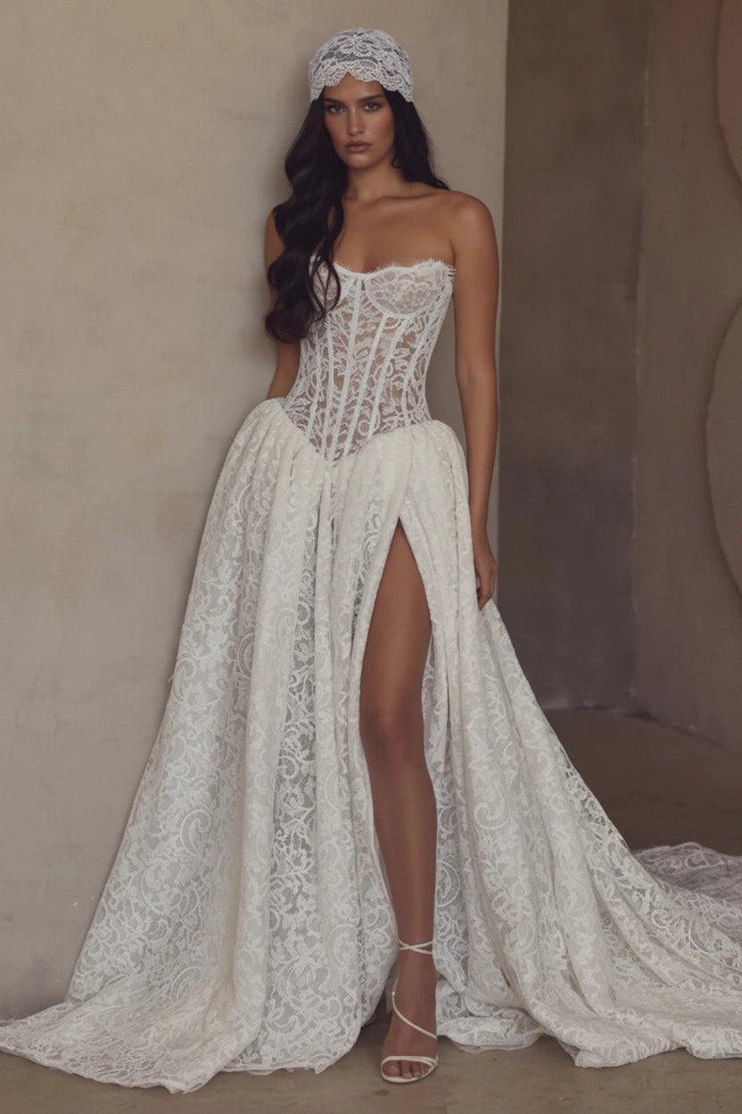 A woman models a strapless white lace wedding gown with a fitted corset-style bodice, high front slit, and long flowing train against a warm beige textured wall.