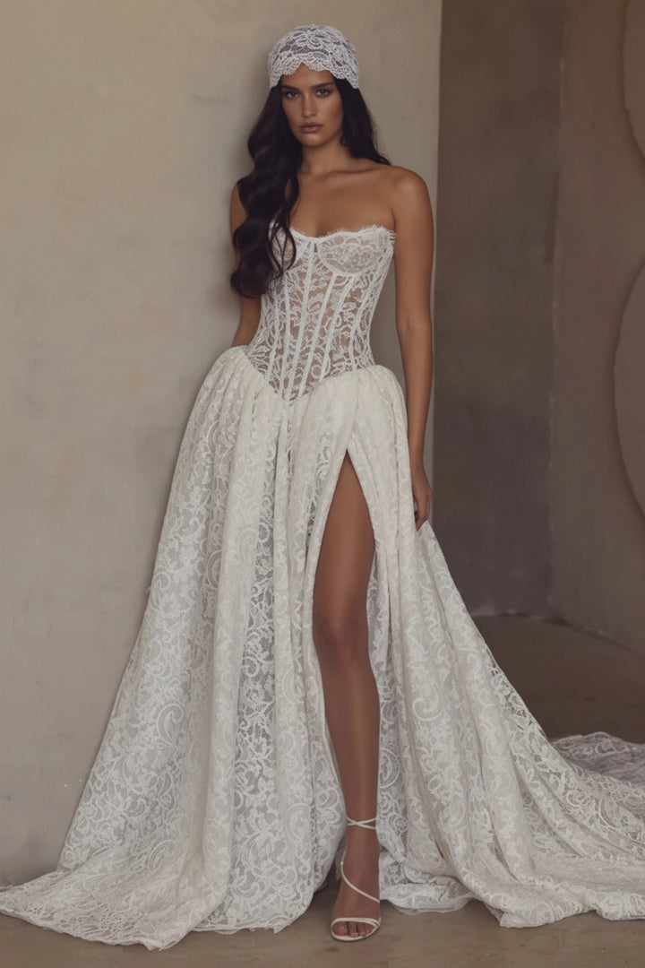 A woman models a strapless white lace wedding gown with a fitted corset-style bodice, high front slit, and long flowing train against a warm beige textured wall.