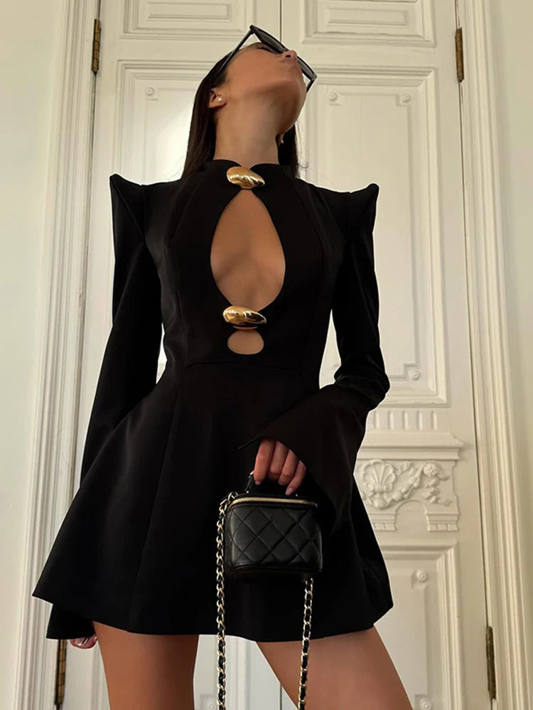 Person wearing a black dress with cut-out design, holding a black handbag against a white wall.