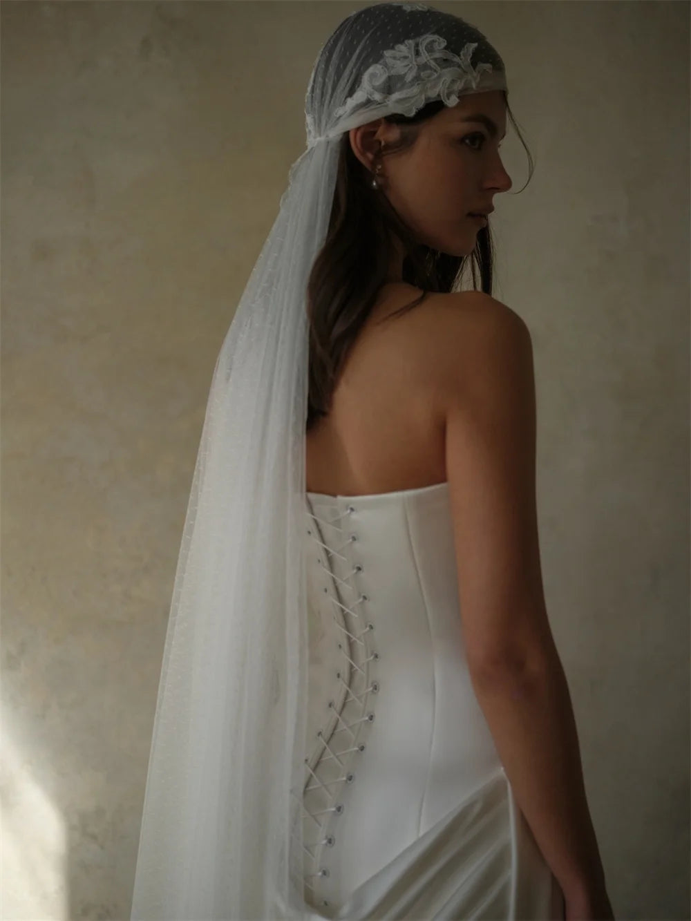A woman in a white strapless wedding dress with lace-up back detailing, wearing a sheer gray veil with white floral embroidery, photographed against a textured beige wall.