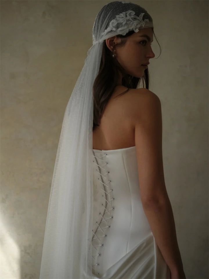 A woman in a white strapless wedding dress with lace-up back detailing, wearing a sheer gray veil with white floral embroidery, photographed against a textured beige wall.