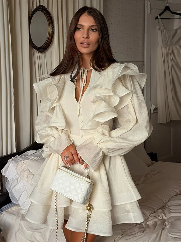 Woman in a white ruffled dress holding a white handbag in a room with curtains and a mirror.